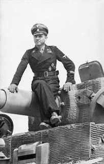man, wearing dress uniform and a cap, sits on top of a tank barrel 